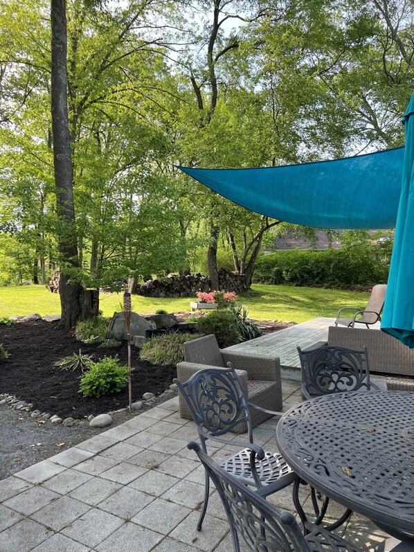 Un espace patio comprenant une table ronde en métal et deux chaises en osier, le tout entouré d’une bordure en pierre et protégé par une voile d’ombrage bleue.