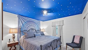 Een slaapkamer met een bed, nachtkastjes, een stoel, een wandtapijt en een plafond met sterrenmotief.