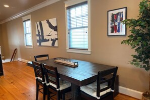 Un área de comedor con una mesa y sillas de madera, dos ventanas con persianas y dos cuadros en la pared.