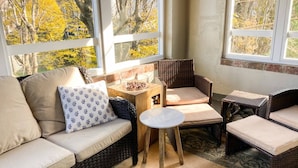 Een gezellig hoekje met een bank, een kleine ronde tafel en rieten stoelen, allemaal gericht op een groot raam met uitzicht op bomen.