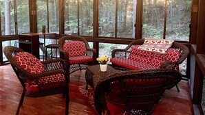 A cozy seating area with wicker chairs and a table, large windows with string lights, and a view of a forest.