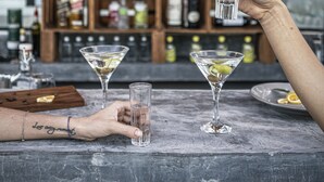 A bar with a wooden shelf stocked with bottles, two martini glasses on the counter, and a person holding a glass. - Clandestino Hotel Recreo - Adults Only (San Miguel de Allende)