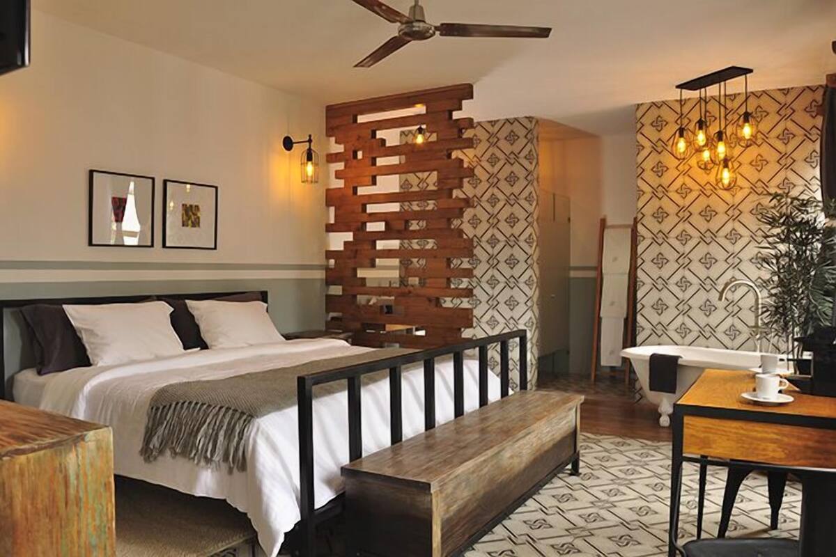 Une chambre à coucher comprenant un lit, un banc en bois, un bureau avec une chaise, un ventilateur de plafond et un mur décoré d’un motif.