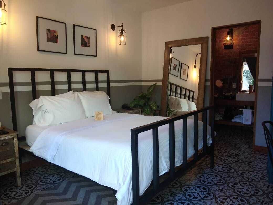 Una cama bien hecha con sábanas blancas, cabecera de madera y mesita de noche con lámpara. En la pared hay cuadros enmarcados y un espejo que refleja la habitación.