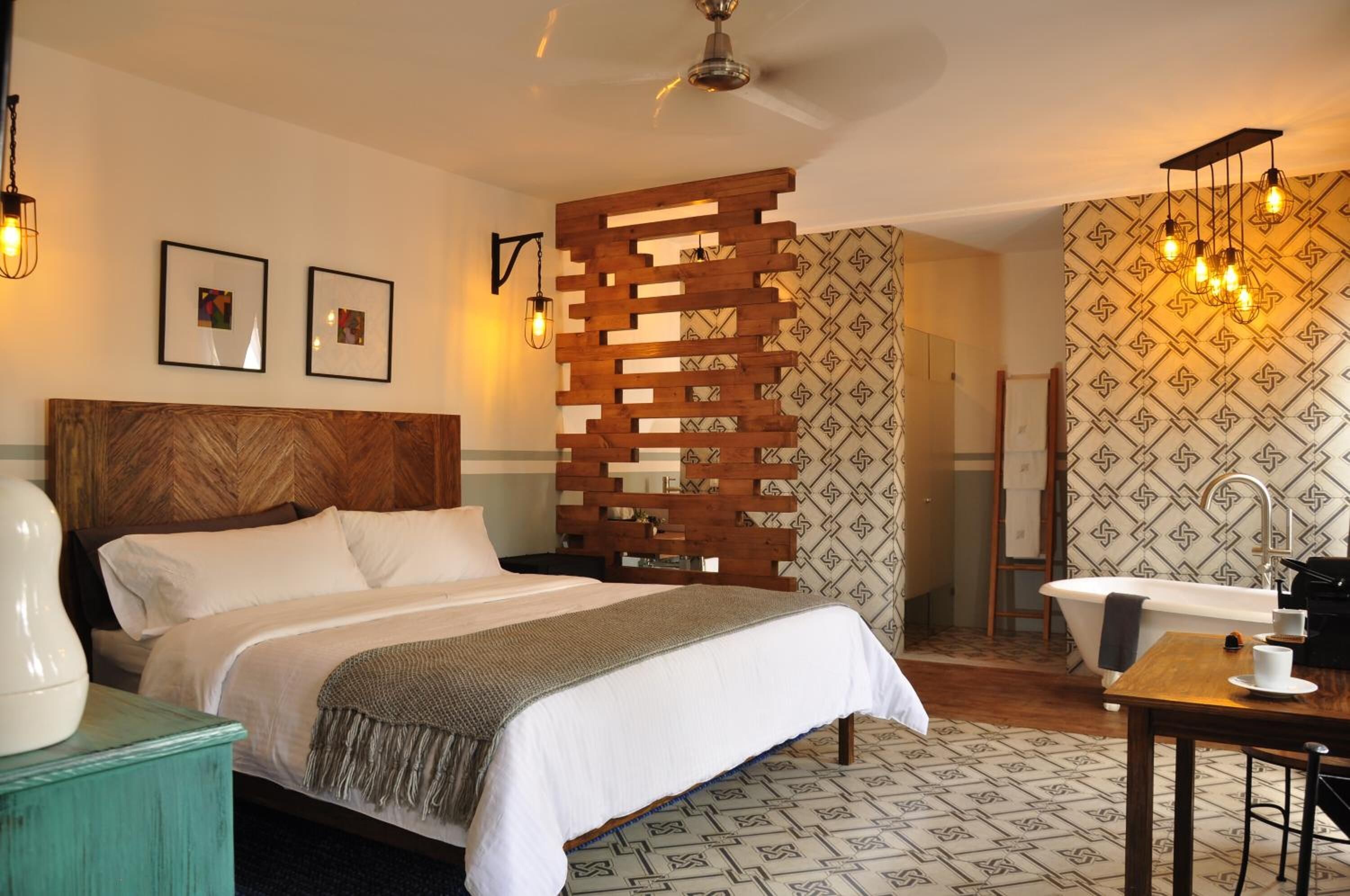 A bedroom with a wooden headboard, a bed with white linens, a wooden partition, a bathroom with a bathtub, and patterned wallpaper.