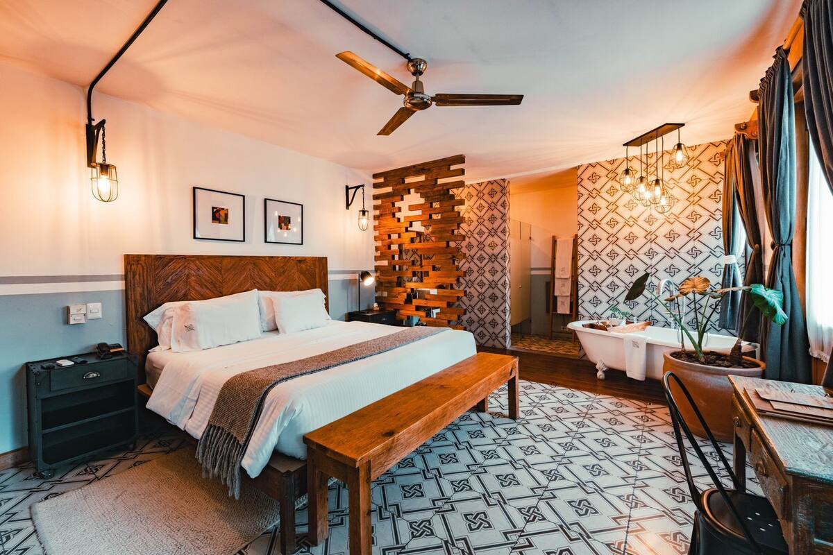 Une chambre d’hôtel moderne avec un grand lit, une tête de lit en bois, un ventilateur de plafond, un panneau mural en bois, un papier peint à motifs, une baignoire blanche et une petite table avec une chaise.