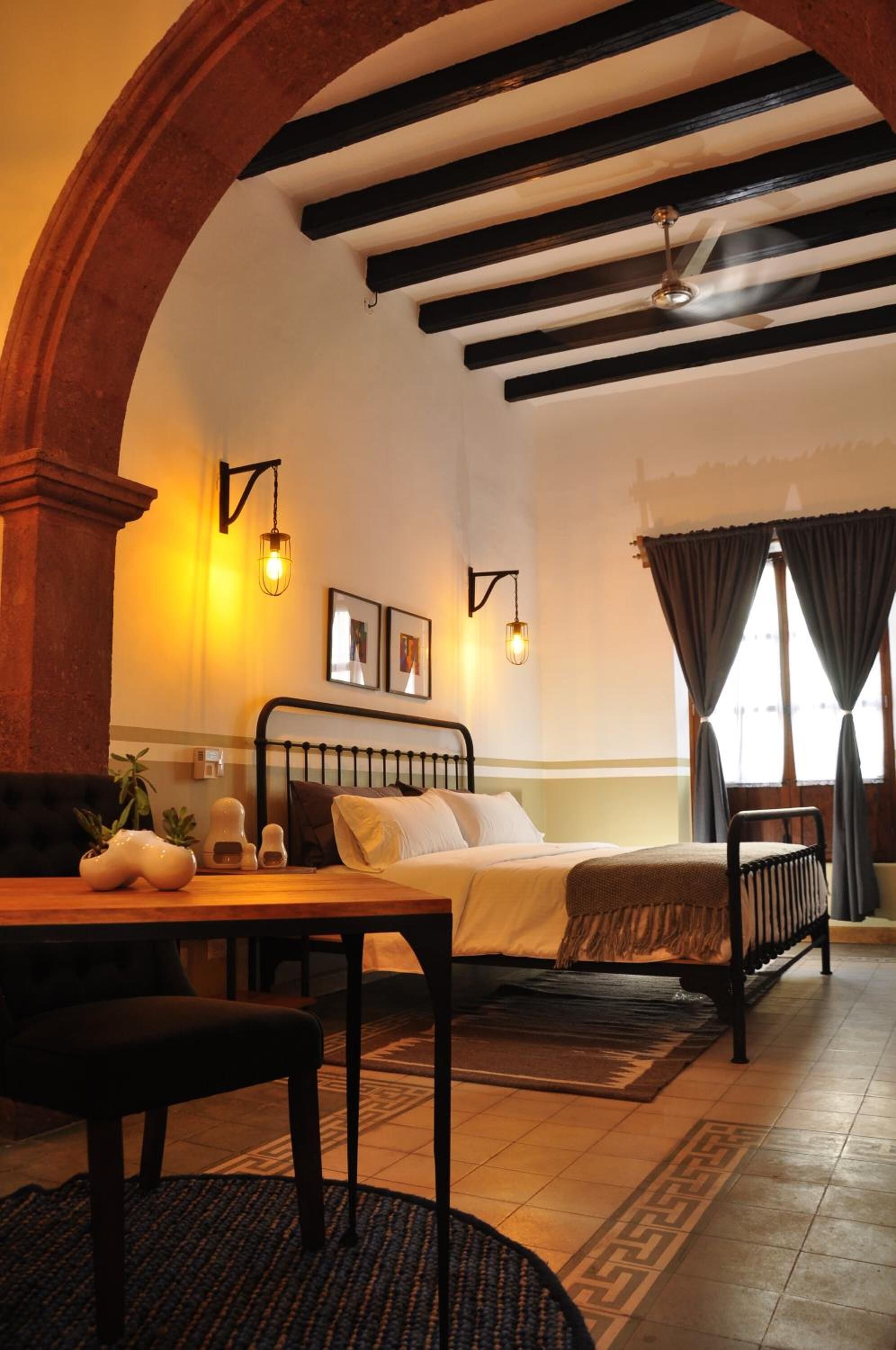 Une pièce confortable avec un lit, une table et des chaises. Il y a une fenêtre avec des rideaux, un luminaire fixé au mur et un ventilateur de plafond.