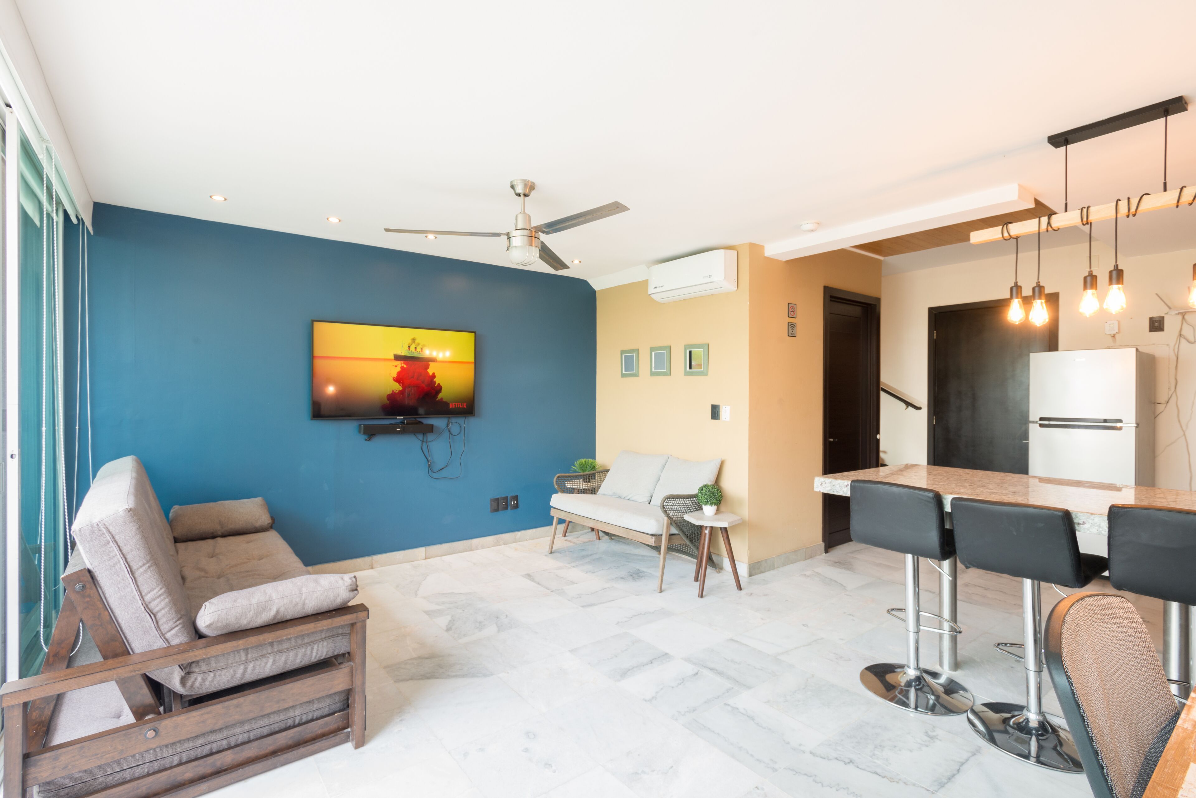 A modern living room with a blue accent wall, a sofa, a small table with a plant, and a kitchen area with a bar and seating.