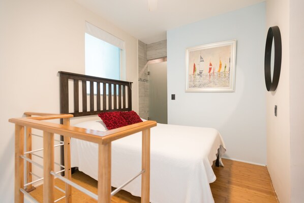 Un dormitorio con una cama, una escalera de madera, un cojín rojo, un cuadro enmarcado y un objeto circular negro en la pared.