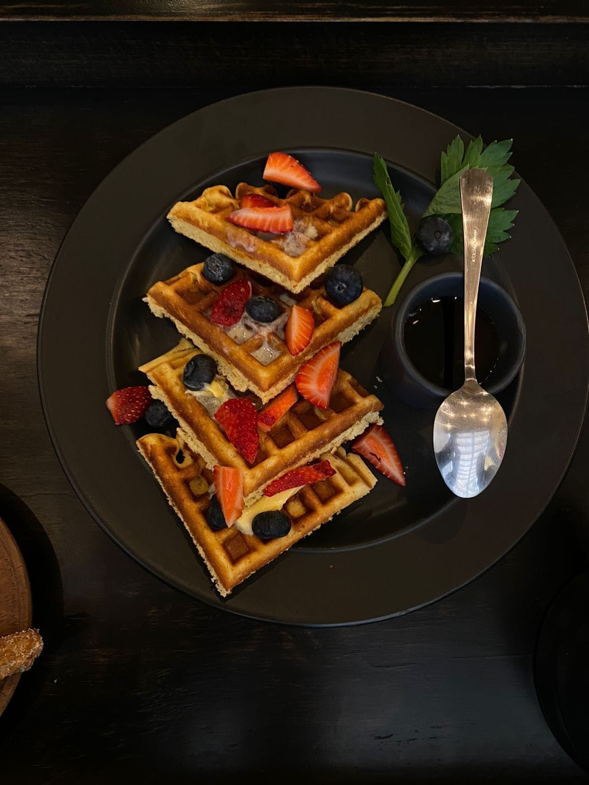 A plate of waffles topped with strawberries and blueberries, accompanied by a spoon and a small bowl of syrup.