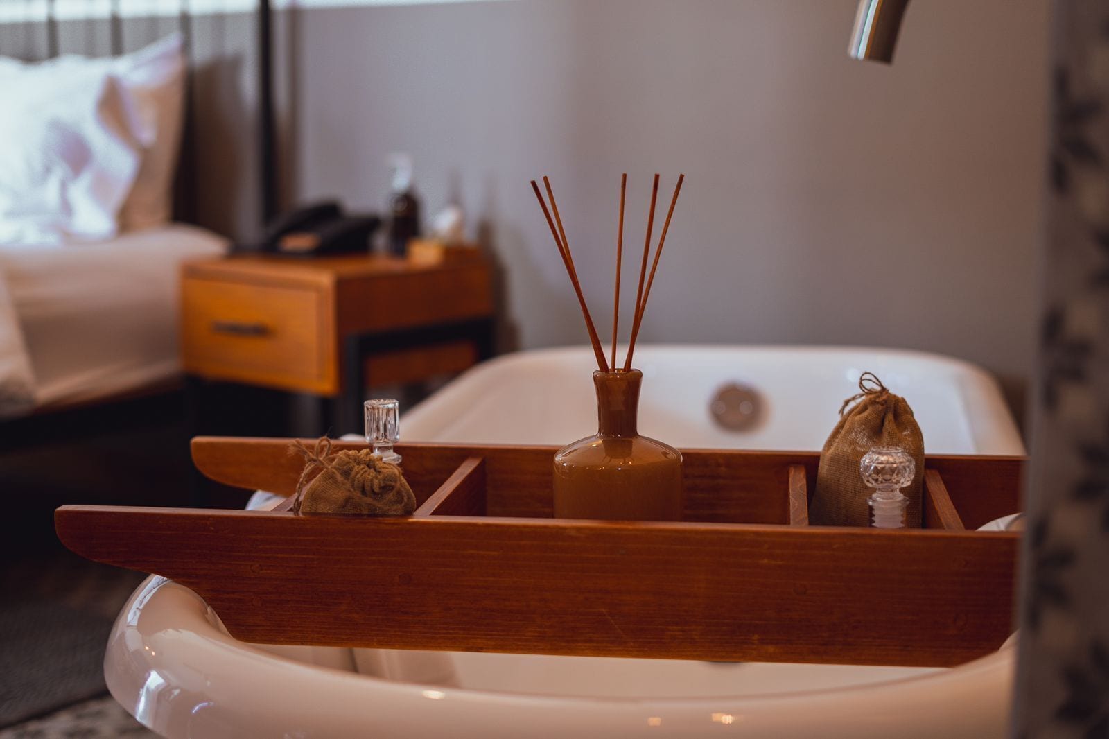 A modern bathroom with a wooden vanity, a white bathtub, and a small bedside table with a lamp.