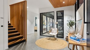 Um interior moderno com uma escada de madeira, um tapete redondo trançado, uma cadeira branca, uma mesa com livros e uma planta, e uma porta de vidro que dá para outro cômodo.