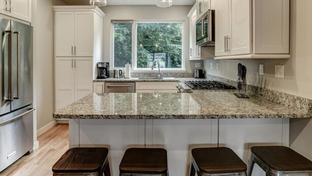 A modern kitchen with white cabinets, granite countertops, a large island with stools, stainless steel appliances, and a window providing a view of trees. - Private Elevator in Hot South Slope (Asheville)