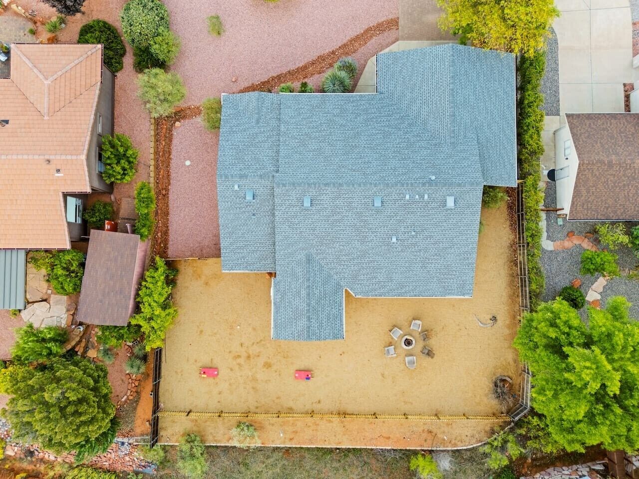 An aerial view of a residential property with a large backyard, a patio area, and surrounding greenery.