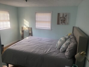 Un dormitorio con una cama, dos ventanas con persianas, un espejo y una obra de arte colgada en la pared.