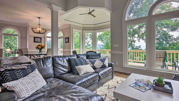 A spacious living room with a leather sofa, large windows, and a ceiling fan. - Spacious Elk Mountain Sanctuary w/ Deck & Views! (Asheville)