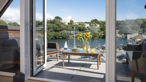 Ein Balkon mit Blick auf einen Fluss, ein Tisch mit einer Vase Sonnenblumen und Gartenmöbel.