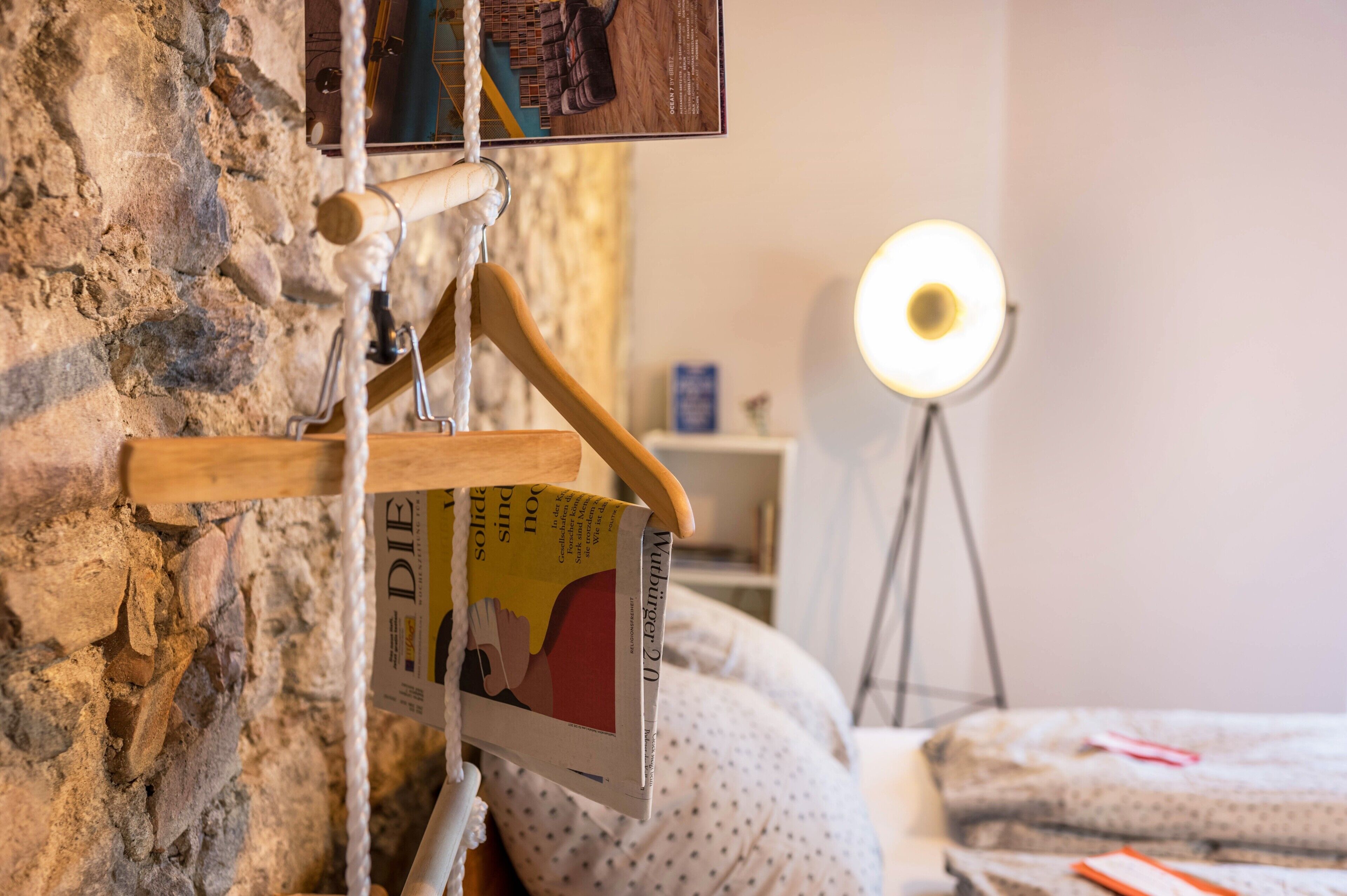 Une pièce avec un mur en pierre, un porte-manteau en bois sur lequel est posé un livre, une lampe de chevet et un lit recouvert d’une couverture à pois.