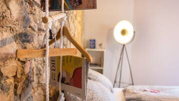 Une pièce avec un mur en pierre, un porte-manteau en bois sur lequel est posé un livre, une lampe de chevet et un lit recouvert d’une couverture à pois.