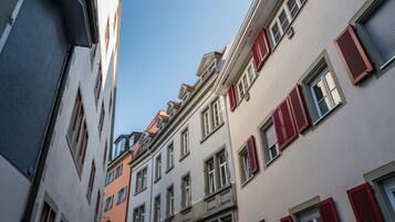 Una calle estrecha flanqueada por edificios de varios pisos, con contraventanas rojas y un cielo azul despejado.