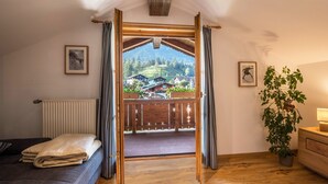 Ein Zimmer mit einem Bett, Holzboden, Heizkörper, Topfpflanze und Blick auf einen Balkon mit Holzgeländer.
