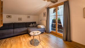 Ein gemütliches Zimmer mit einem dunkelgrauen Sofa, einem kleinen runden Tisch mit Kerzen, Holzdielen und einem großen Fenster mit Vorhängen.