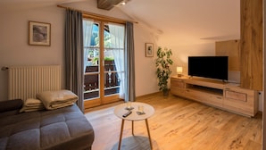 Ein Wohnzimmer mit einer Couch, einem Fernseher auf einem Holzschrank, einem kleinen Tisch mit Gläsern und einem Fenster mit Vorhängen.