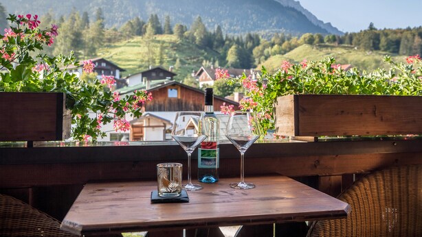 Ein Holztisch mit zwei Weingläsern und einer Flasche, vor einer Berglandschaft und Häusern.