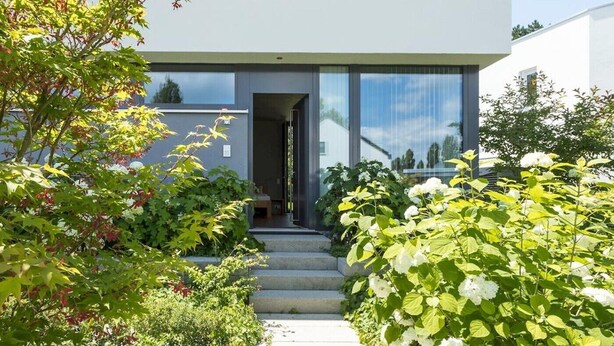 A modern building with a clear entrance, surrounded by lush greenery and white flowers.