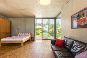 Ein Schlafzimmer mit einem Bett, einem Holzschrank, einer schwarzen Ledersofa und einem großen Fenster mit Blick auf Grünflächen.