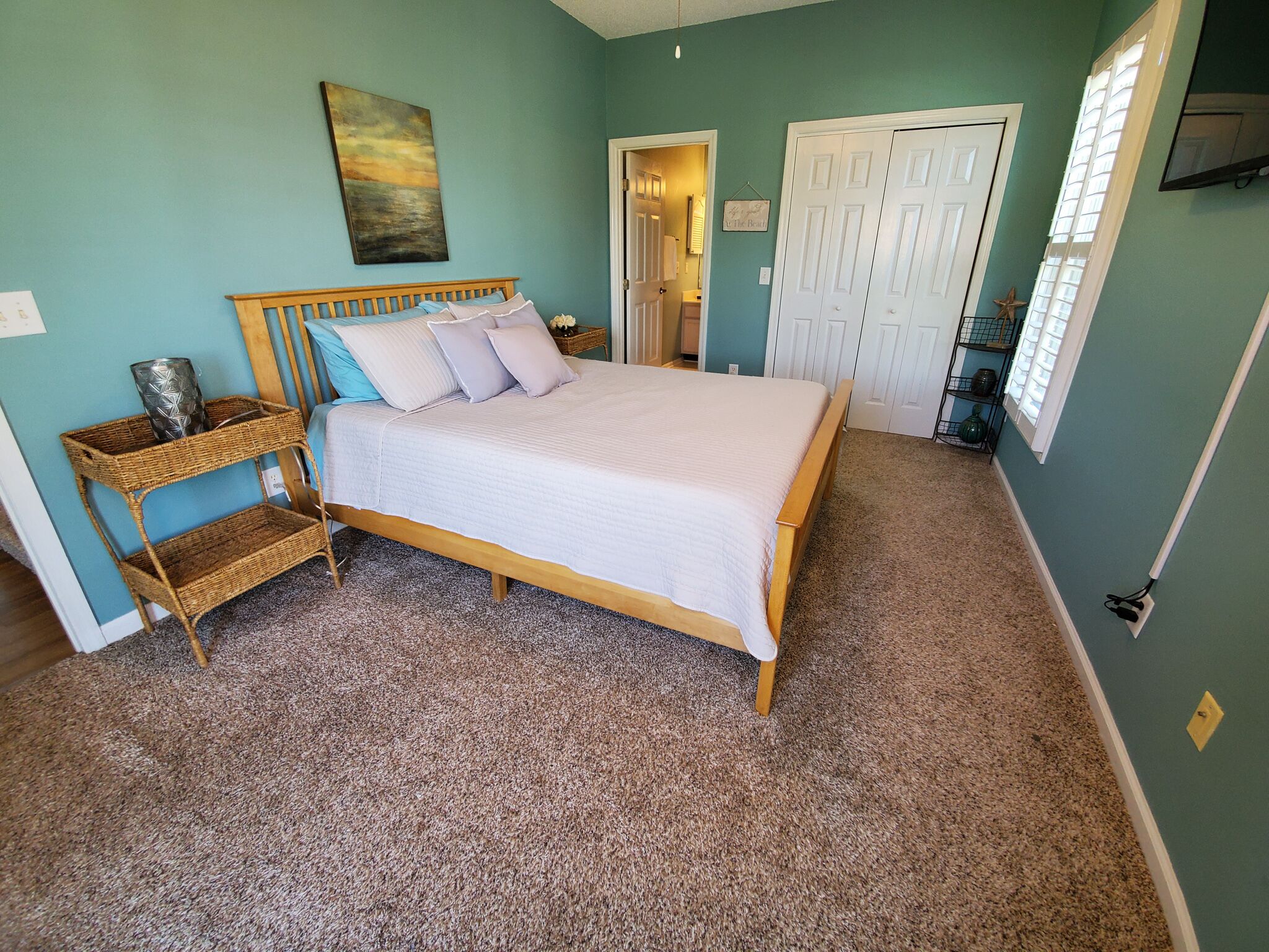 Un dormitorio con cama de madera, mesita de mimbre, un cuadro en la pared y una puerta que da a otra habitación.