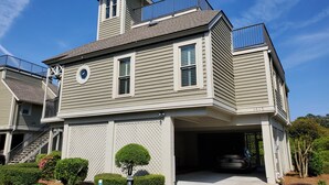 Une maison à deux étages avec un garage, un petit balcon à l’étage supérieur et un jardin bien entretenu à l’avant, agrémenté de haies et d’un arbre.
