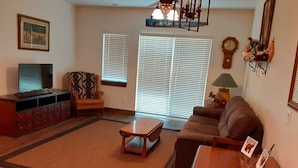 Una sala de estar con una mesa de centro de madera, un sofá, un televisor sobre un mueble bajo y un ventilador de techo.