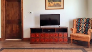 Una sala de estar con una puerta de madera, un televisor sobre un mueble de madera, un sillón estampado y un cuadro en la pared.