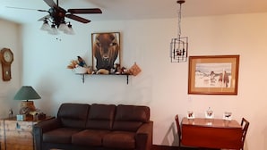 Una sala de estar con un sofá marrón, una mesa de centro de madera, una mesa de comedor con sillas, un ventilador de techo, estantes empotrados con objetos decorativos y un cuadro en la pared.
