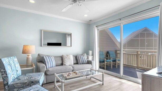 Amplia sala de estar con sofá gris, sillón azul con estampado floral y mesa de centro de vidrio. Se ve un ventilador de techo y un ventanal con vistas a una terraza de madera.