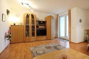 Ein geräumiges Wohnzimmer mit Holzböden, einem großen Schrank mit Glastüren, einem Fernseher und einer Glastür zu einem Balkon.