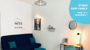Ett modernt vardagsrum med en svart soffa, turkosa kuddar, ett litet turkost bord, ett trägolv, en geometrisk matta, ett konstverk på väggen, en hylla med dekorativa föremål, ett skrivbord med en stol och en golvlampa.