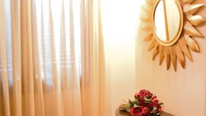 A room with a curtained window, a wooden bench, a small table with a mirror and a bouquet of flowers.
