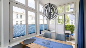 Een lichte eetruimte met een houten tafel, stoelen en een blauwe tafelkleed. Grote ramen bieden uitzicht op gebouwen en een blauwe hekwerk.