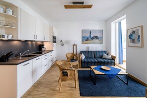 A modern kitchen with white cabinets, a built-in oven, and a coffee machine. There is a dining area with a blue rug, a wooden table, and wicker chairs. A sofa with patterned cushions is against the wall, and a painting of a lighthouse is visible.