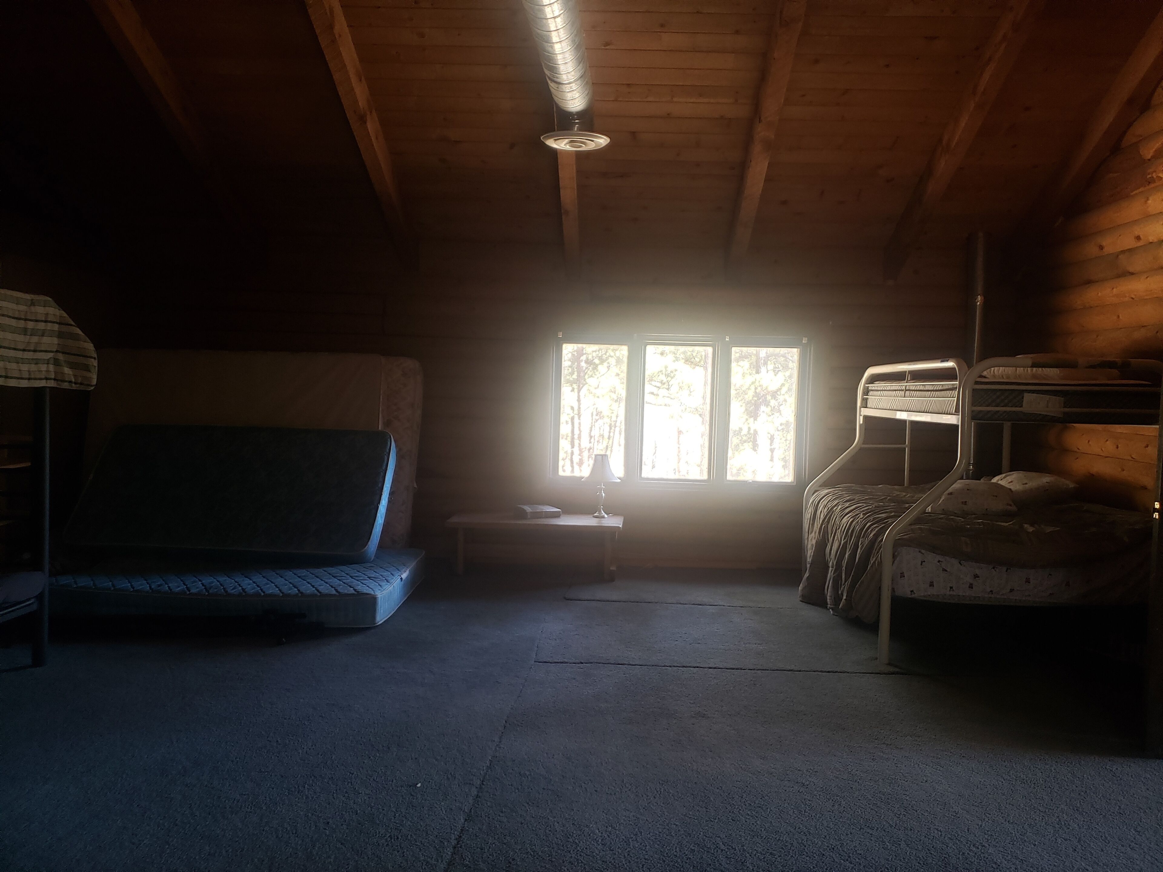 Een kamer in gedimd licht met een stapelbed, een enkel bed en een bureau met een lamp. Drie ramen zorgen voor daglicht.