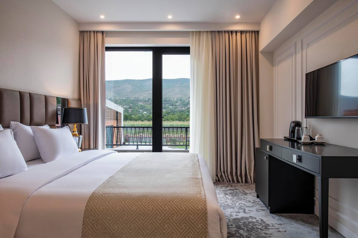 A hotel room with a large bed, a desk, a TV, and a balcony with a view of the mountains.