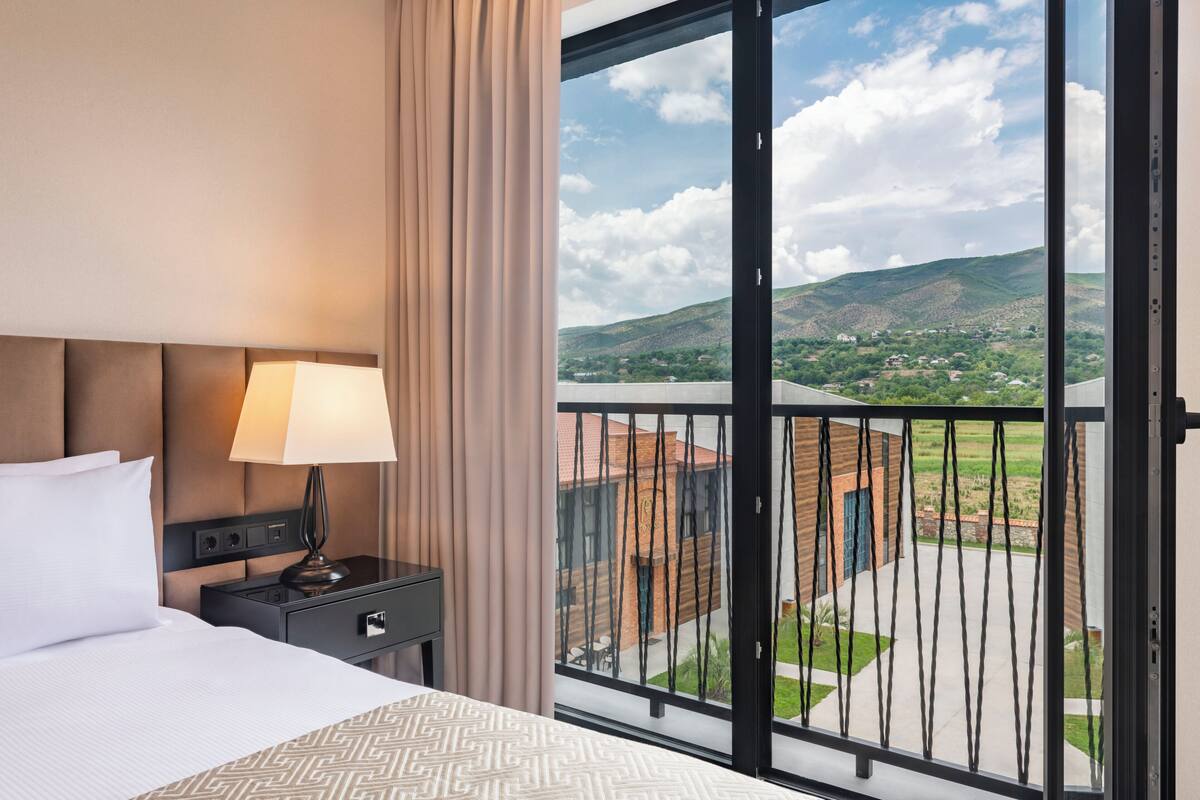 A hotel room with a bed, a nightstand, a lamp, and a view of a balcony with a landscape.
