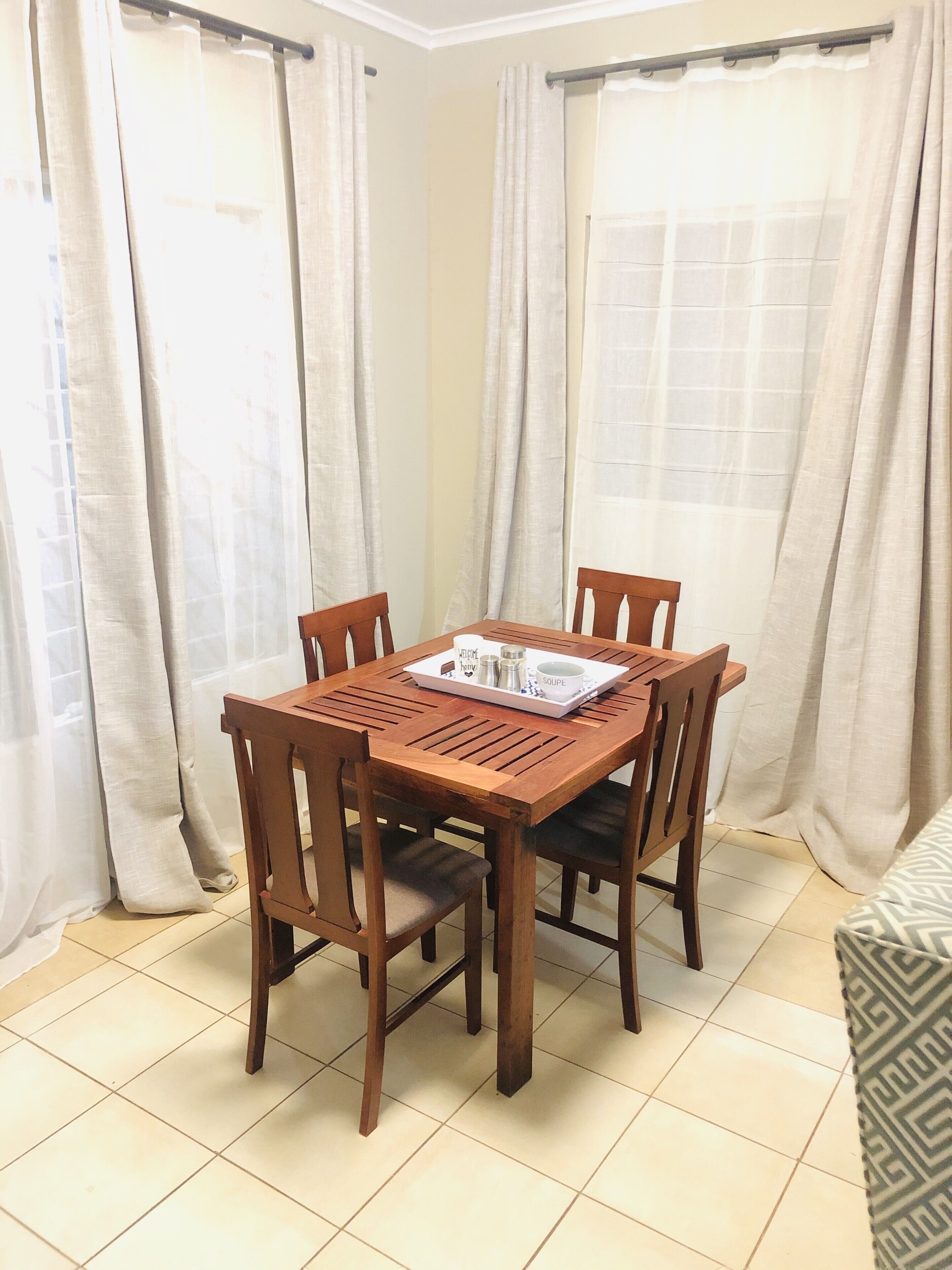 A dining area with a wooden table and chairs, a tray with condiments, and light-colored curtains.