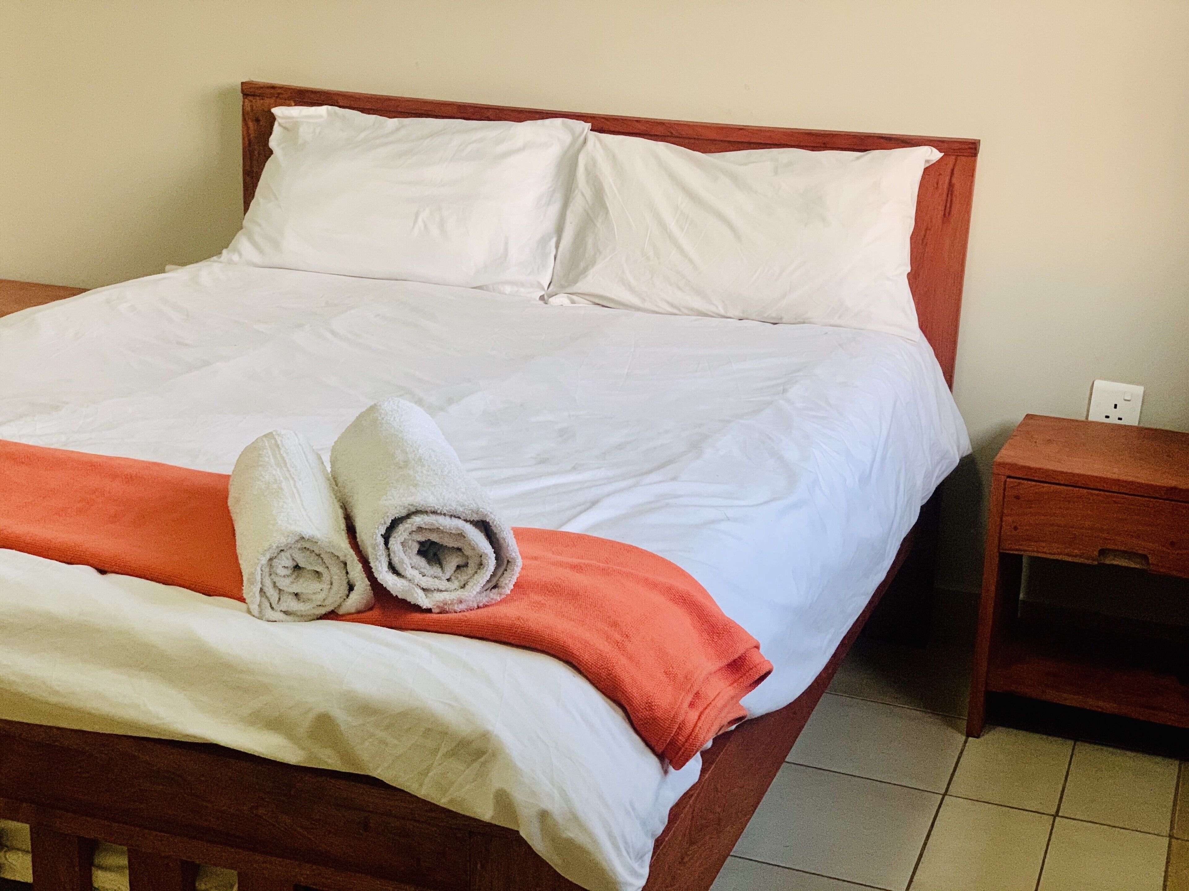 A neatly made bed with white linens and two rolled towels, a wooden headboard, and a small wooden nightstand with a power outlet.