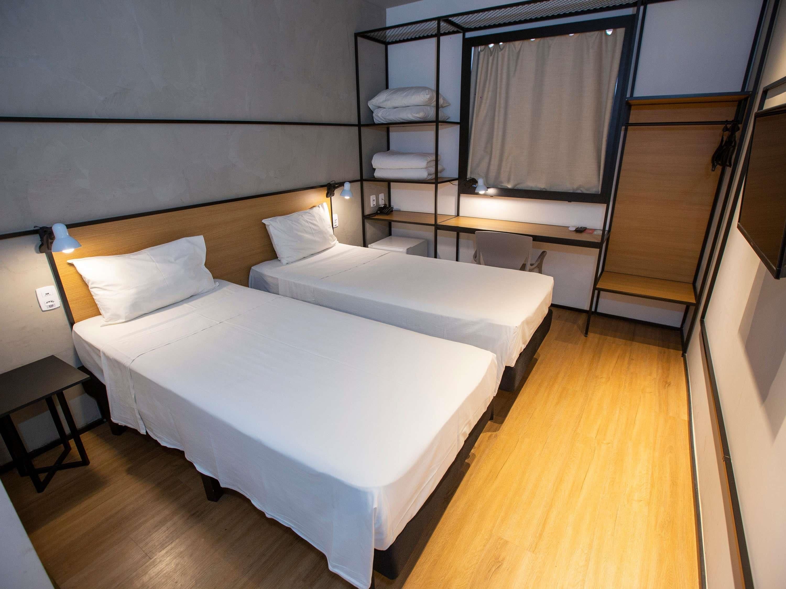 A hotel room with two beds, a desk, a chair, a shelf with towels, and a window with curtains.