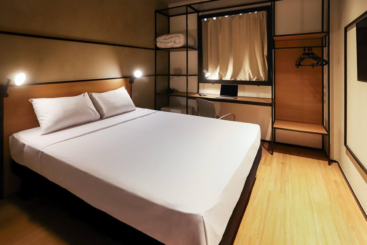 Una cama bien tendida con cabecera, un escritorio con una laptop y un perchero.