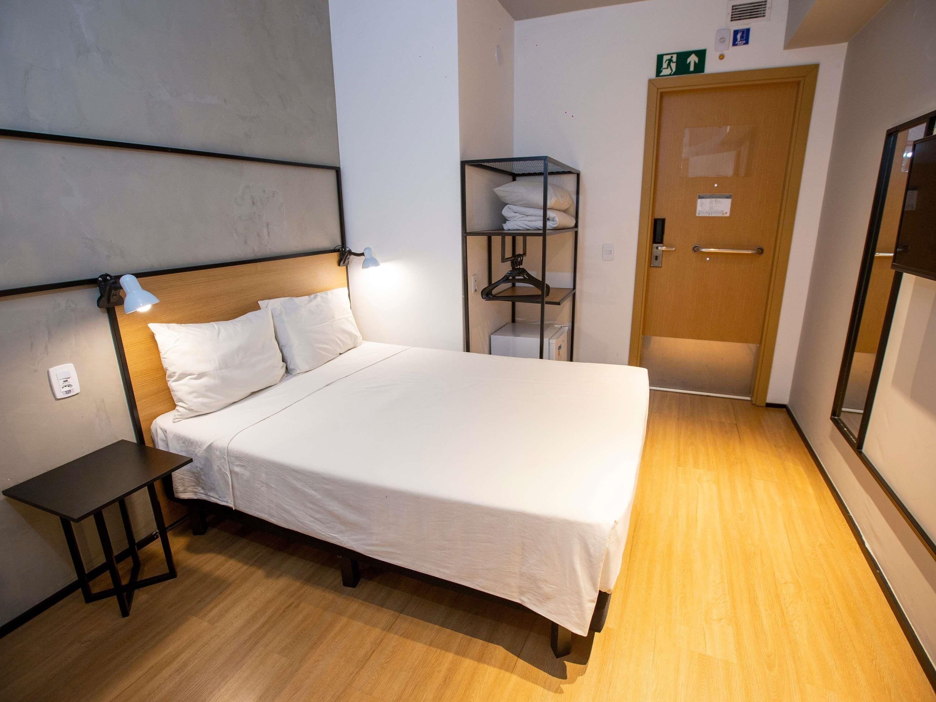 Una habitación de hotel con cama, mesita de noche, lámpara y suelo de madera.