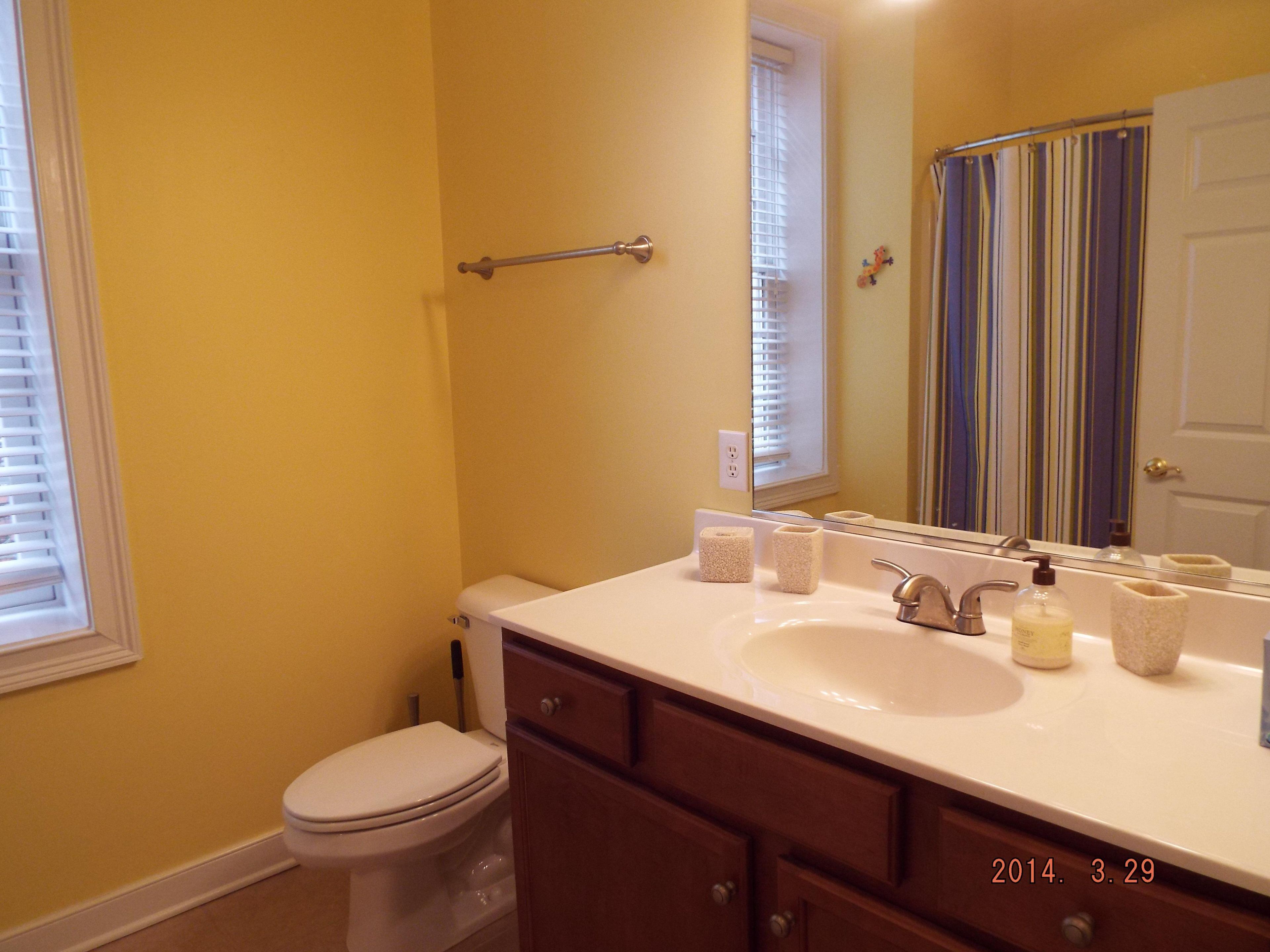 A bathroom with a white sink, a mirror, and a toilet. There is a towel rack above the sink and a window with blinds.